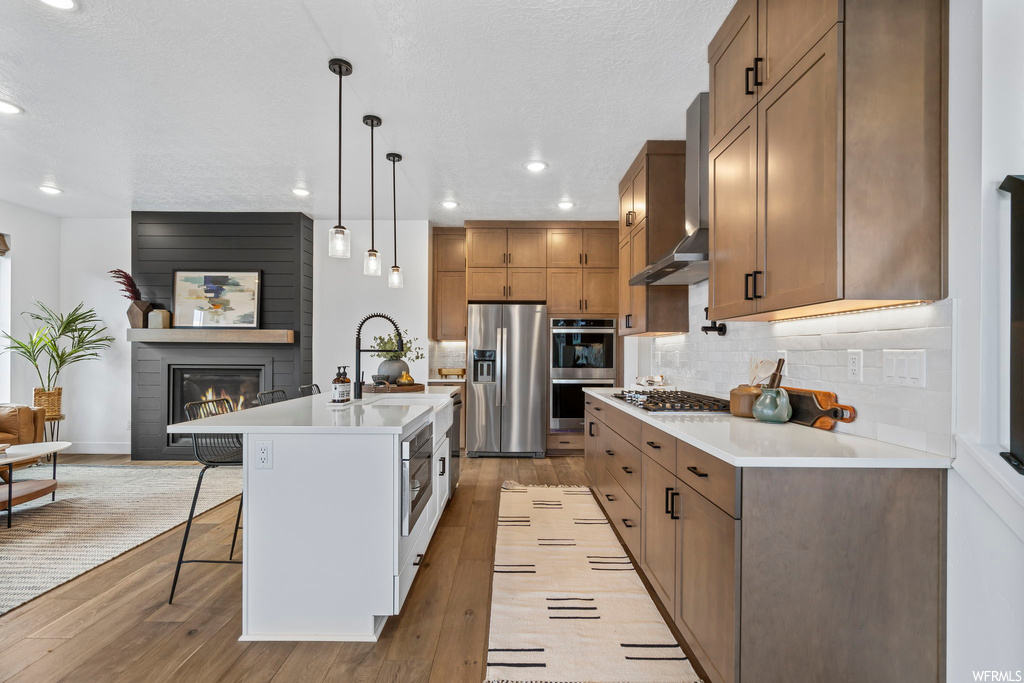 Kitchen featuring light hardwood / wood-style flooring, backsplash, hanging light fixtures, stainless steel appliances, and a kitchen island with sink