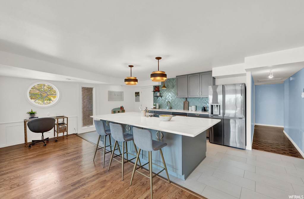 Kitchen featuring gray cabinets, tasteful backsplash, light wood-type flooring, stainless steel refrigerator with ice dispenser, and a kitchen island with sink