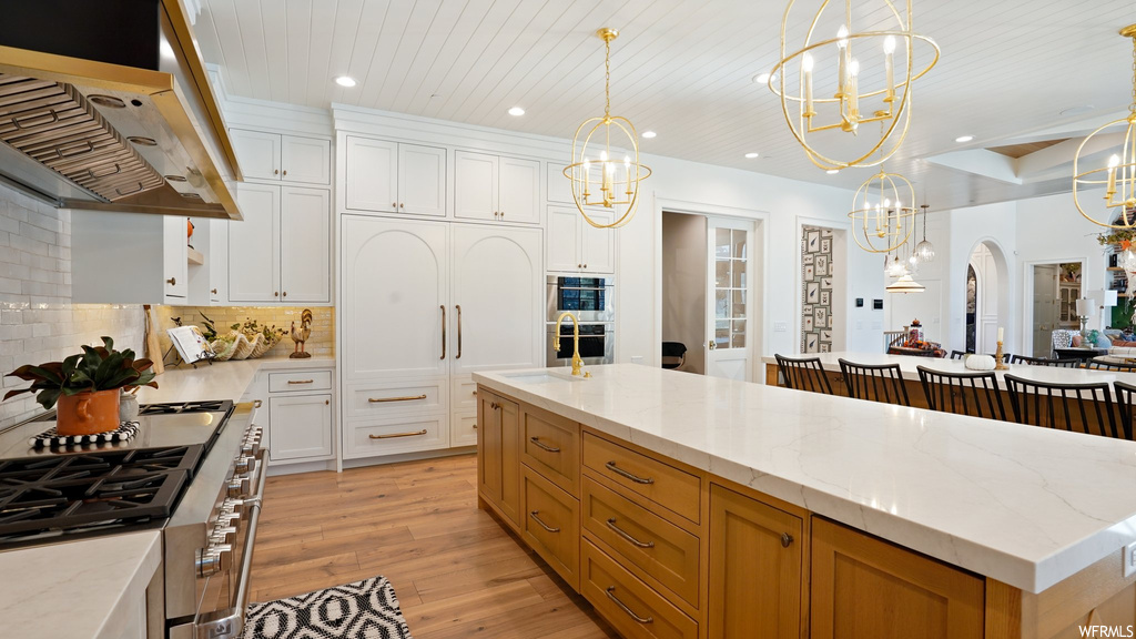Kitchen featuring decorative light fixtures, a center island with sink, light hardwood / wood-style flooring, white cabinets, and wall chimney exhaust hood