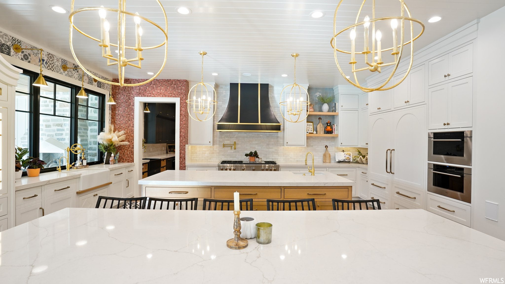 Kitchen featuring pendant lighting, white cabinets, wall chimney range hood, and a notable chandelier
