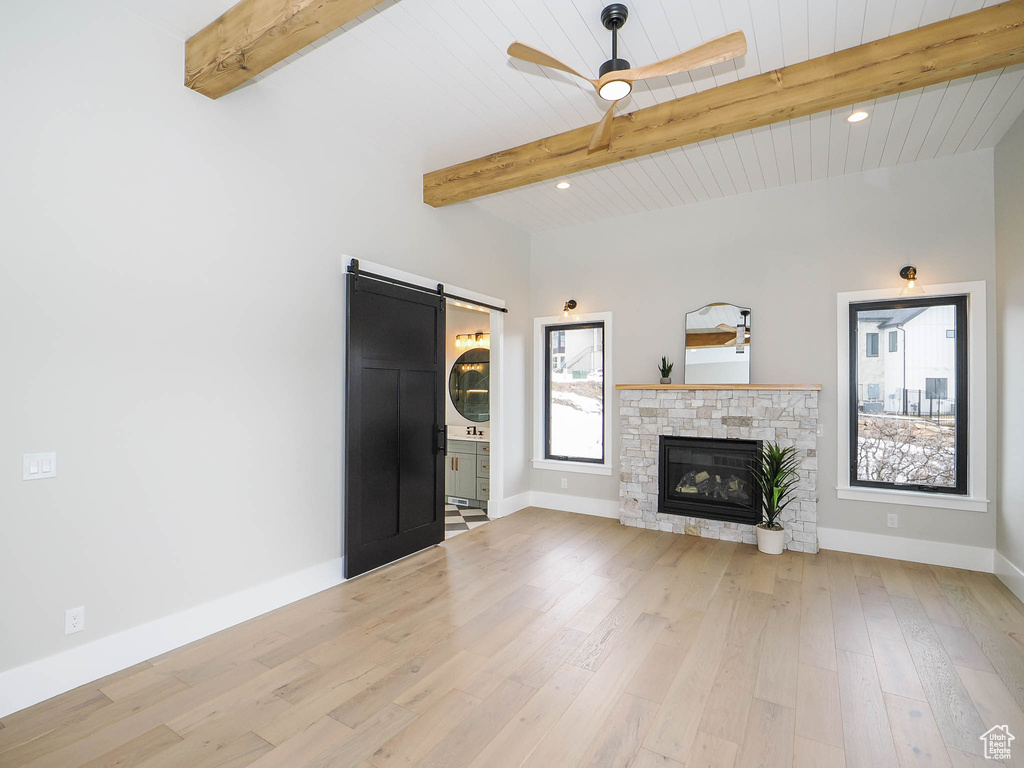 Unfurnished living room featuring light hardwood / wood-style floors, ceiling fan, a barn door, beam ceiling, and a stone fireplace