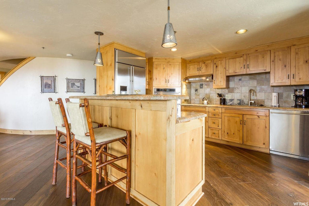 Kitchen featuring decorative light fixtures, dark wood-type flooring, a kitchen island, wall chimney range hood, and backsplash