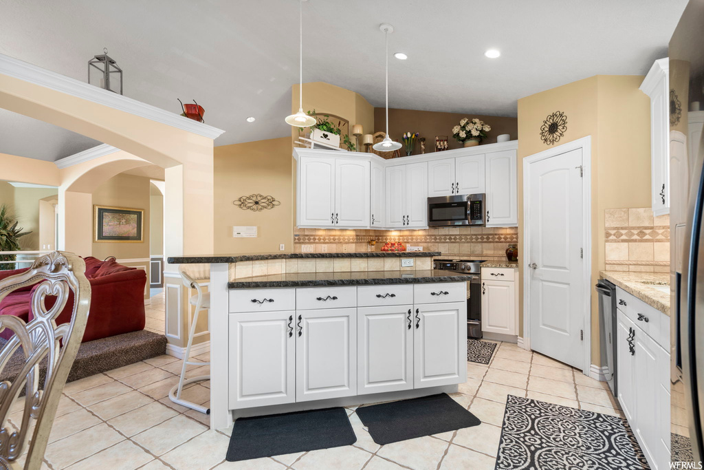 Kitchen featuring tasteful backsplash, dark stone counters, white cabinets, light tile floors, and decorative light fixtures