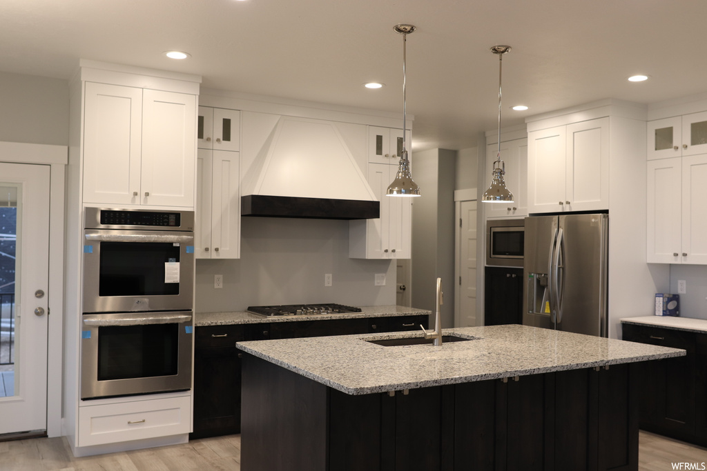Kitchen with premium range hood, sink, light wood-type flooring, a center island with sink, and white cabinets