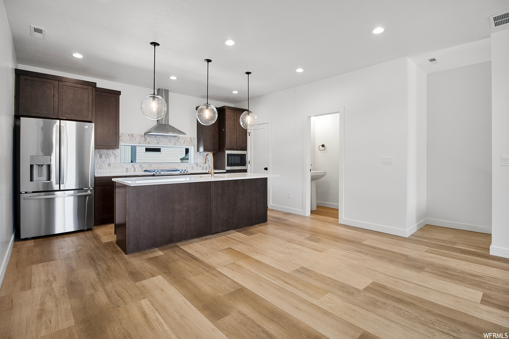 Kitchen with light hardwood / wood-style floors, an island with sink, backsplash, pendant lighting, and wall chimney range hood