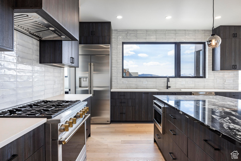 Kitchen with hanging light fixtures, premium appliances, backsplash, wall chimney range hood, and light hardwood / wood-style floors