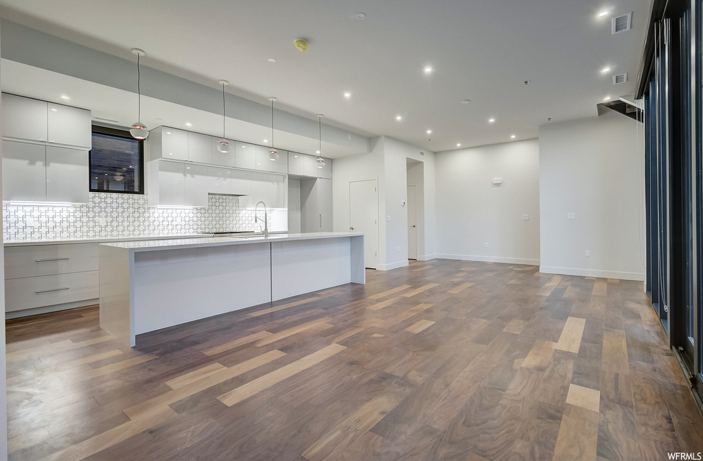 Kitchen featuring dark hardwood / wood-style floors, an island with sink, backsplash, white cabinets, and pendant lighting