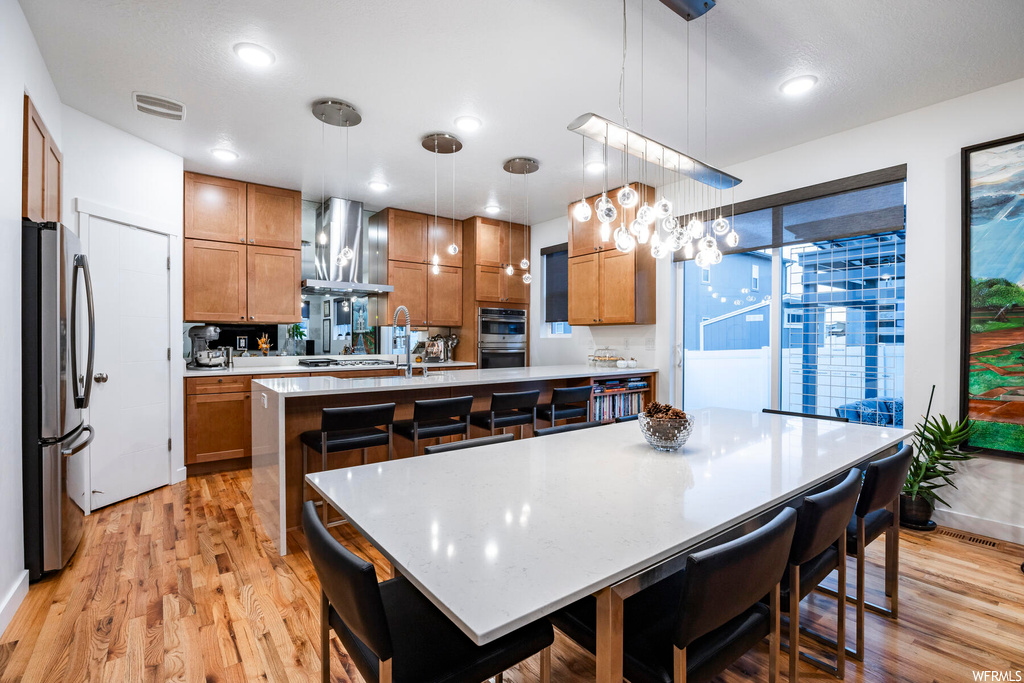 Kitchen featuring light hardwood / wood-style floors, an island with sink, wall chimney range hood, hanging light fixtures, and stainless steel appliances