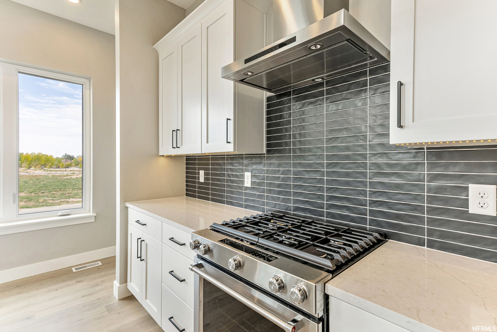 Kitchen with a wealth of natural light, wall chimney range hood, stainless steel range, white cabinets, and backsplash