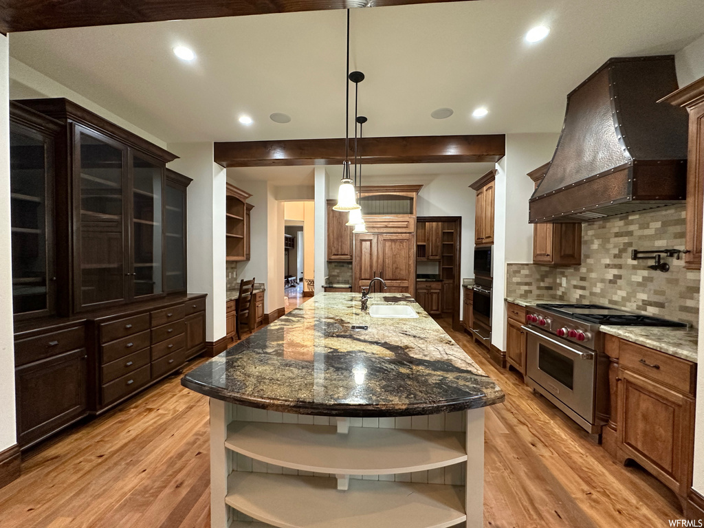 Kitchen featuring premium range hood, dark stone counters, a center island with sink, designer stove, and backsplash