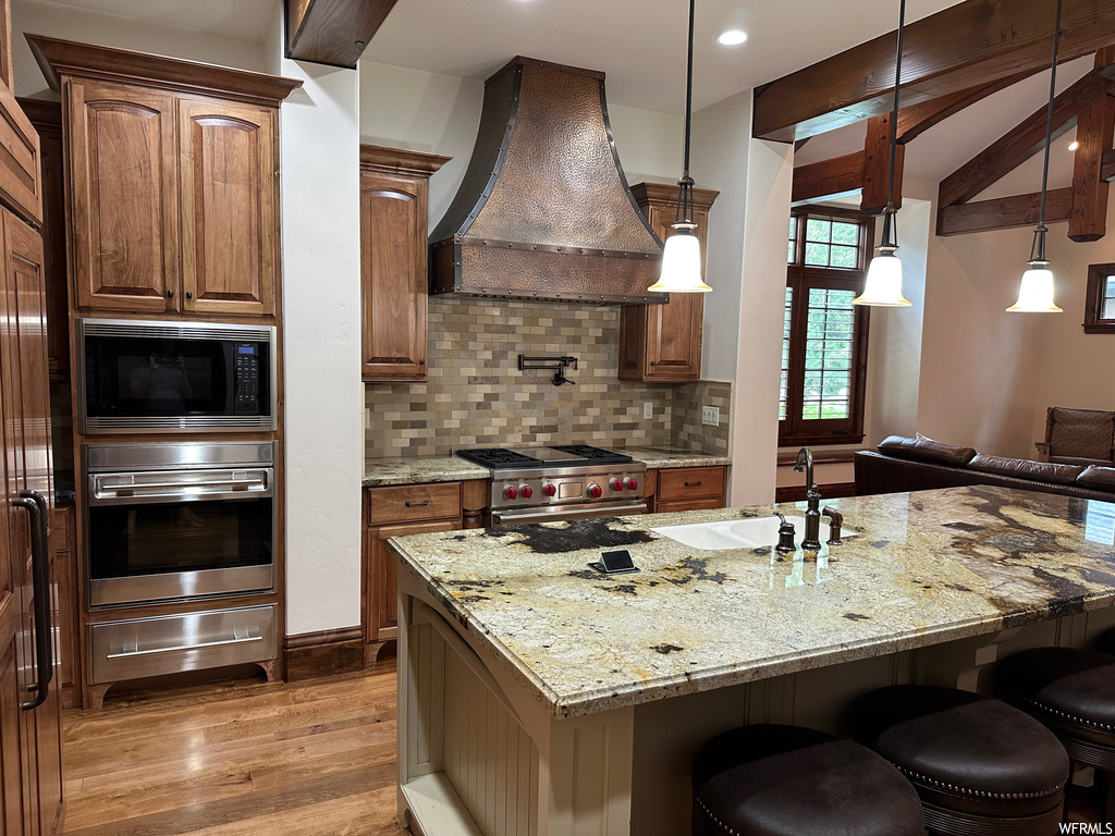 Kitchen with premium range hood, appliances with stainless steel finishes, light stone countertops, light wood-type flooring, and tasteful backsplash