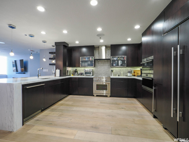 Kitchen with appliances with stainless steel finishes, wall chimney range hood, decorative light fixtures, and light hardwood / wood-style flooring