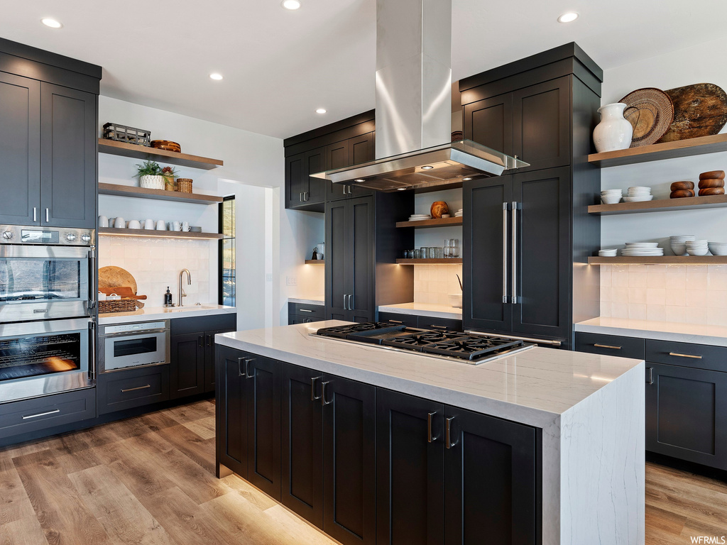 Kitchen with island exhaust hood, light stone counters, appliances with stainless steel finishes, light hardwood / wood-style floors, and tasteful backsplash