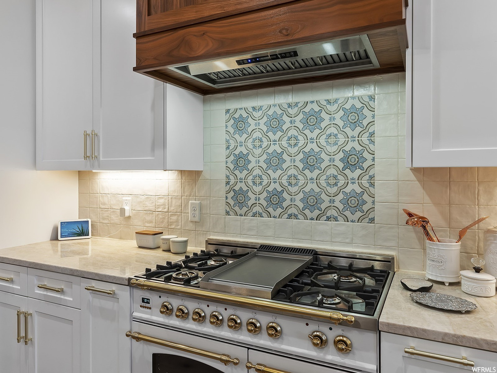 Kitchen with custom range hood, tasteful backsplash, double oven range, light stone counters, and white cabinetry