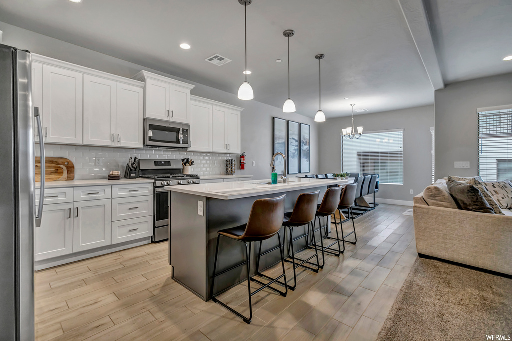 Kitchen featuring a chandelier, a center island with sink, stainless steel appliances, tasteful backsplash, and white cabinetry