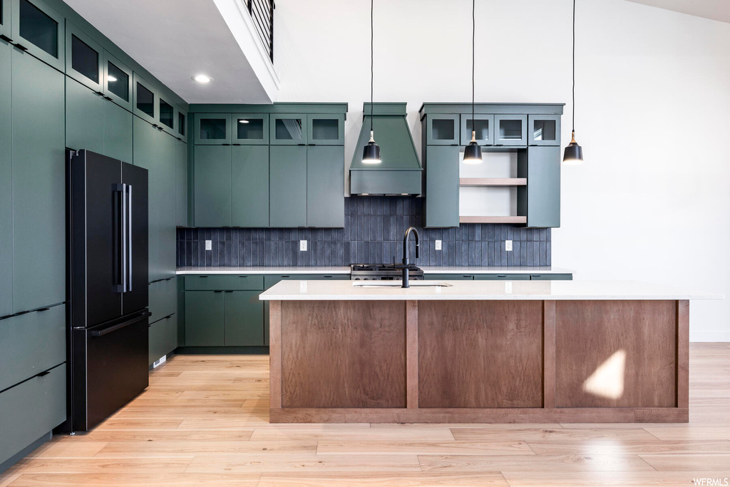 Kitchen featuring custom range hood, a kitchen island with sink, hanging light fixtures, light hardwood / wood-style floors, and high end black refrigerator
