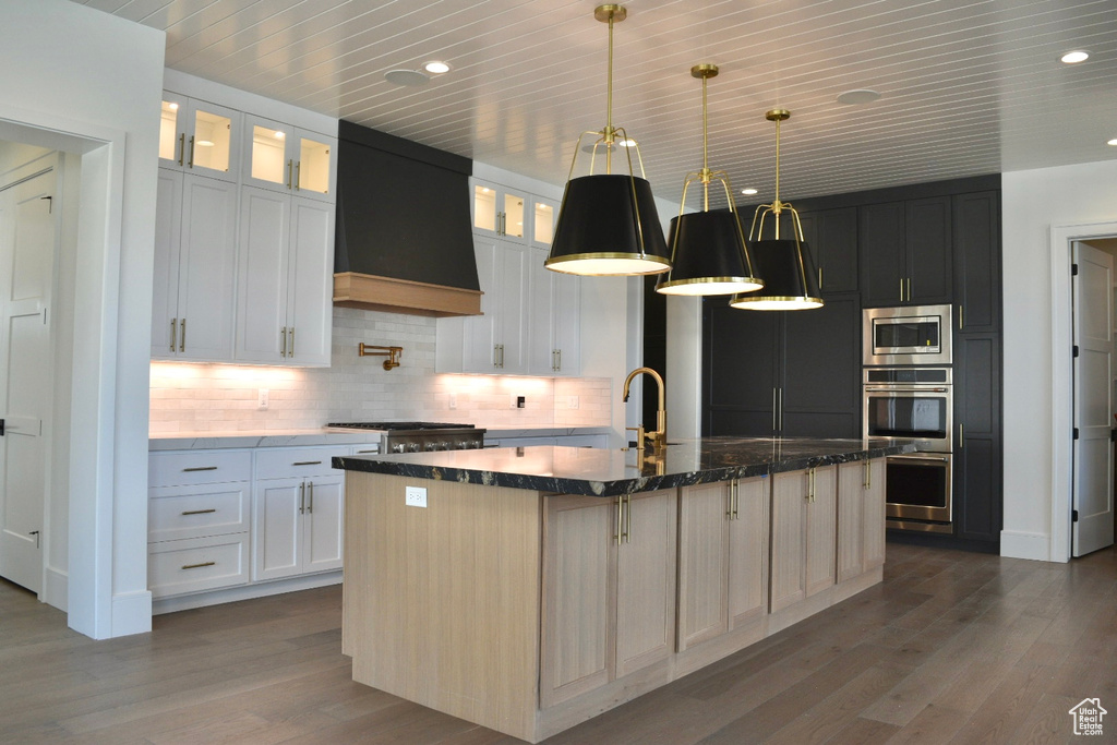 Kitchen with tasteful backsplash, a kitchen island with sink, custom exhaust hood, hardwood / wood-style floors, and hanging light fixtures