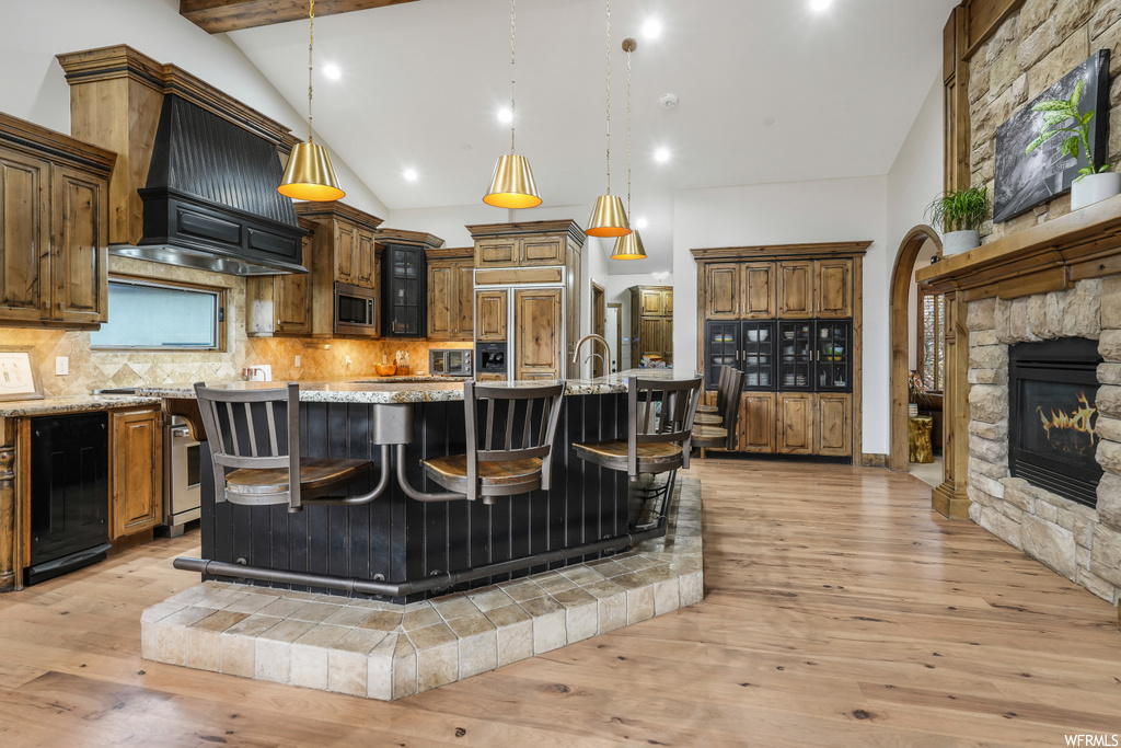 Kitchen with a fireplace, a kitchen island with sink, premium range hood, and light wood-type flooring