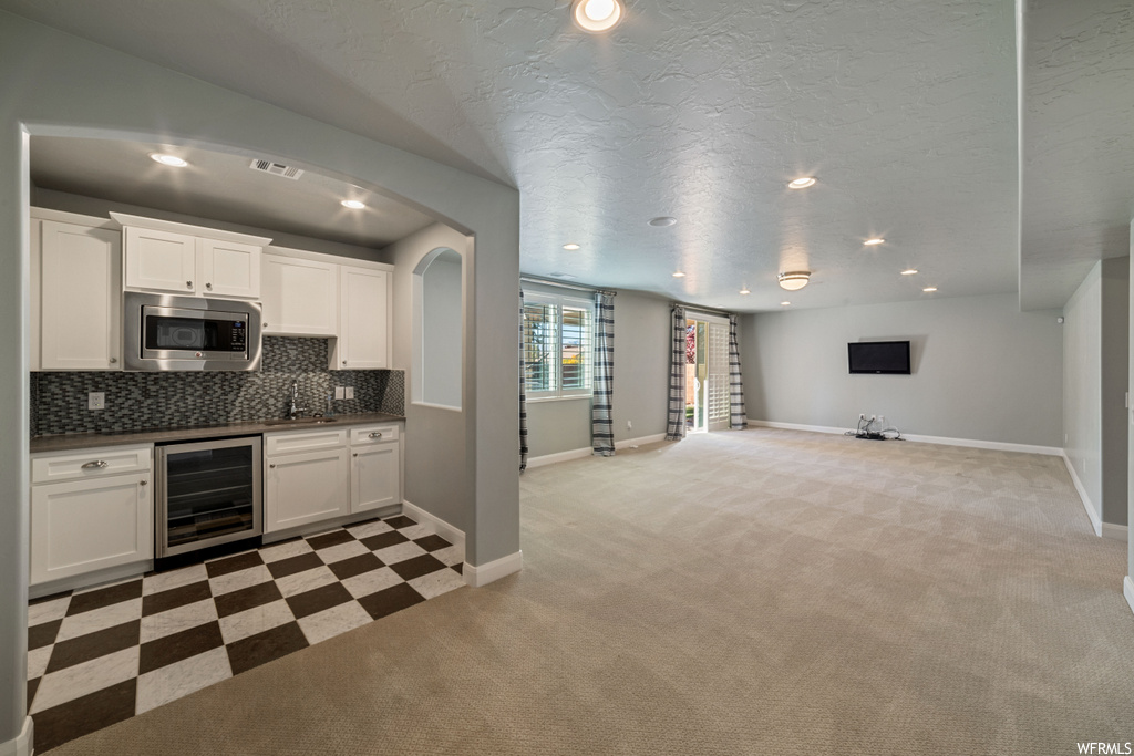 Kitchen featuring stainless steel microwave, beverage cooler, light colored carpet, tasteful backsplash, and white cabinetry