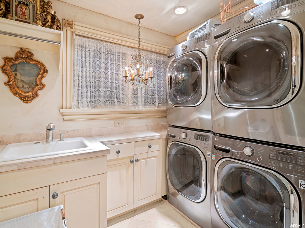 Clothes washing area with cabinets, sink, a chandelier, stacked washing maching and dryer, and light tile floors
