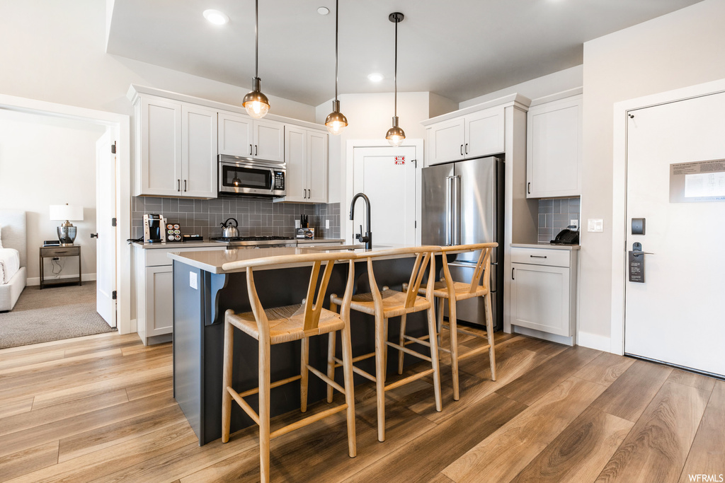 Kitchen with pendant lighting, a kitchen island with sink, stainless steel appliances, backsplash, and light wood-type flooring