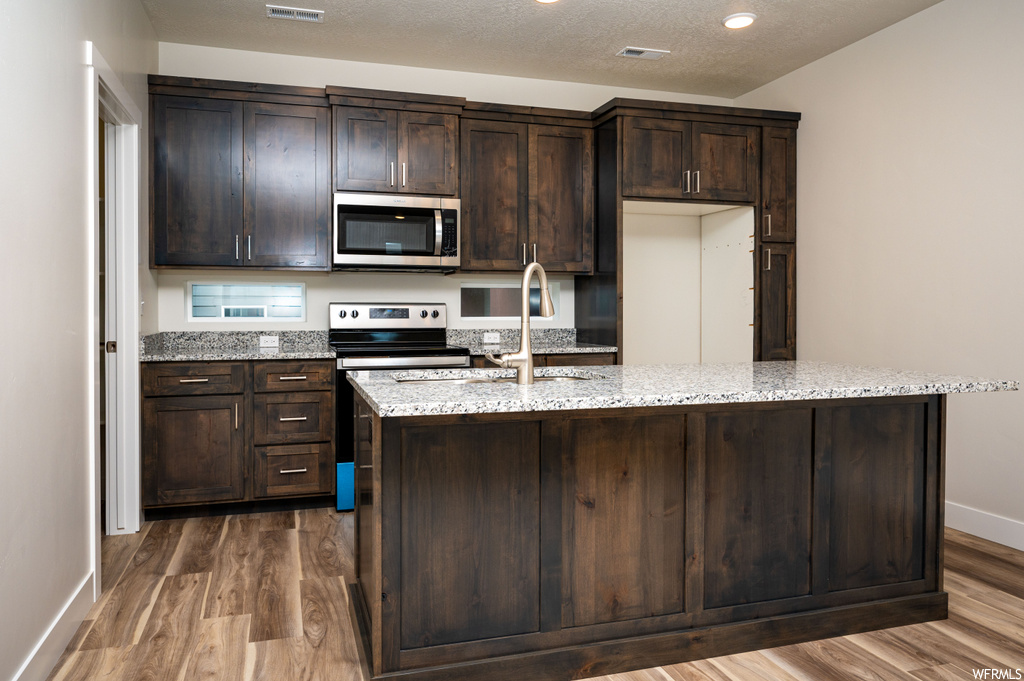 Kitchen featuring hardwood / wood-style flooring, dark brown cabinets, light stone countertops, stainless steel appliances, and a center island with sink