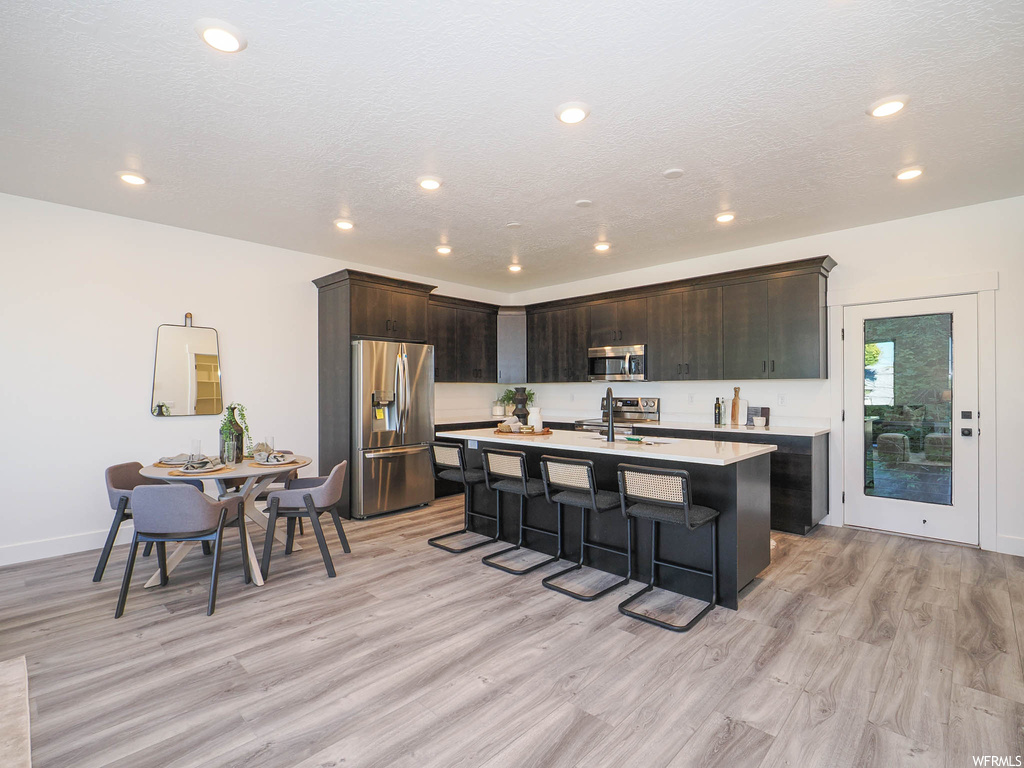 Kitchen featuring an island with sink, appliances with stainless steel finishes, a kitchen bar, dark brown cabinetry, and light hardwood / wood-style flooring