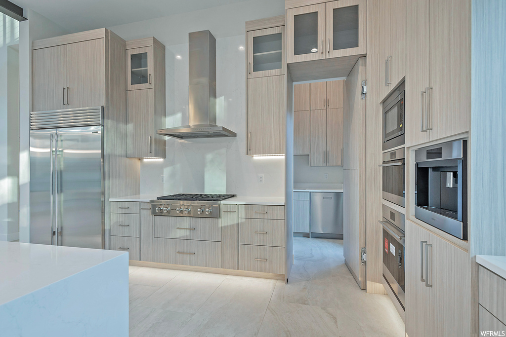 Kitchen featuring light brown cabinets, built in appliances, and wall chimney exhaust hood