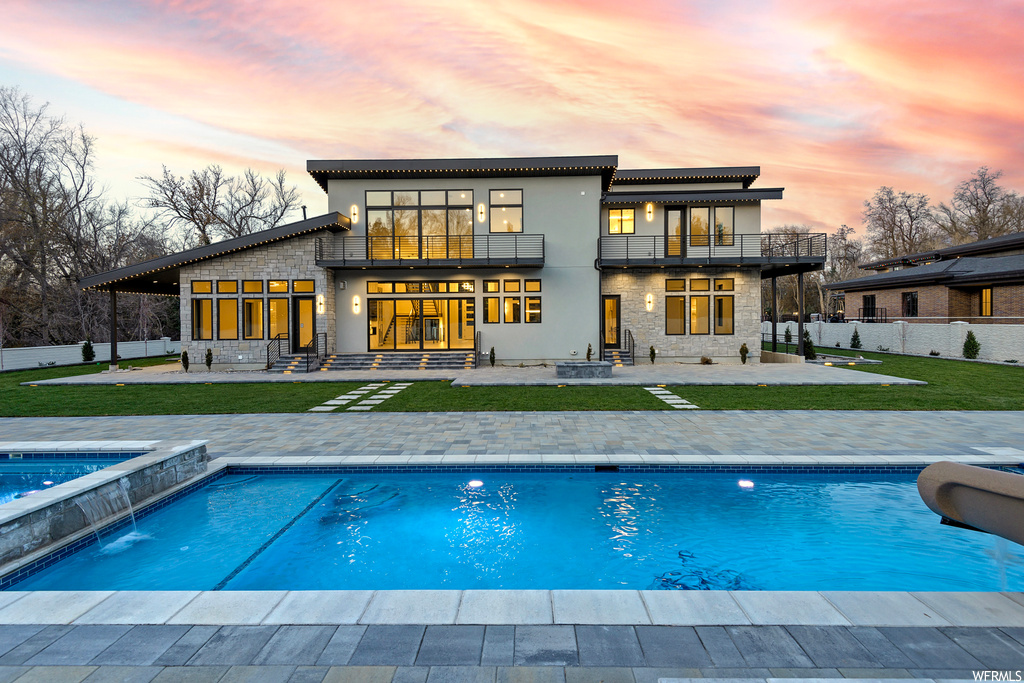Back house at dusk with a swimming pool with hot tub, pool water feature, a patio, and a balcony