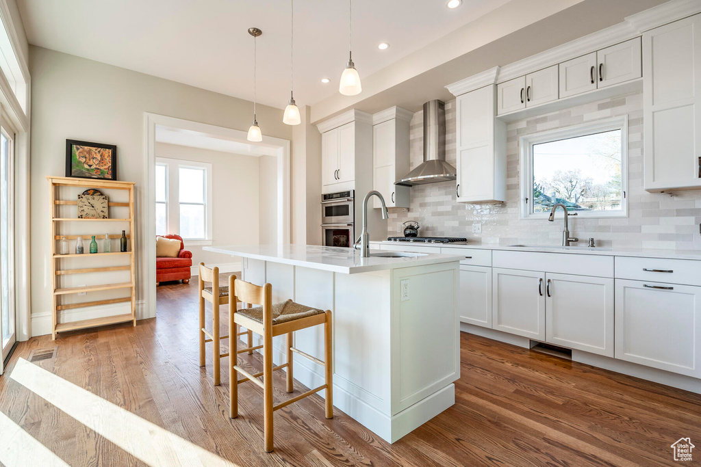 Kitchen featuring wall chimney exhaust hood, an island with sink, and white cabinets