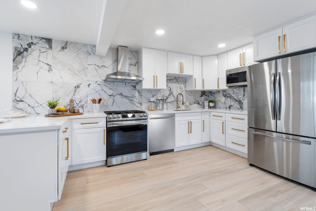 Kitchen featuring light hardwood / wood-style flooring, appliances with stainless steel finishes, wall chimney exhaust hood, white cabinets, and tasteful backsplash