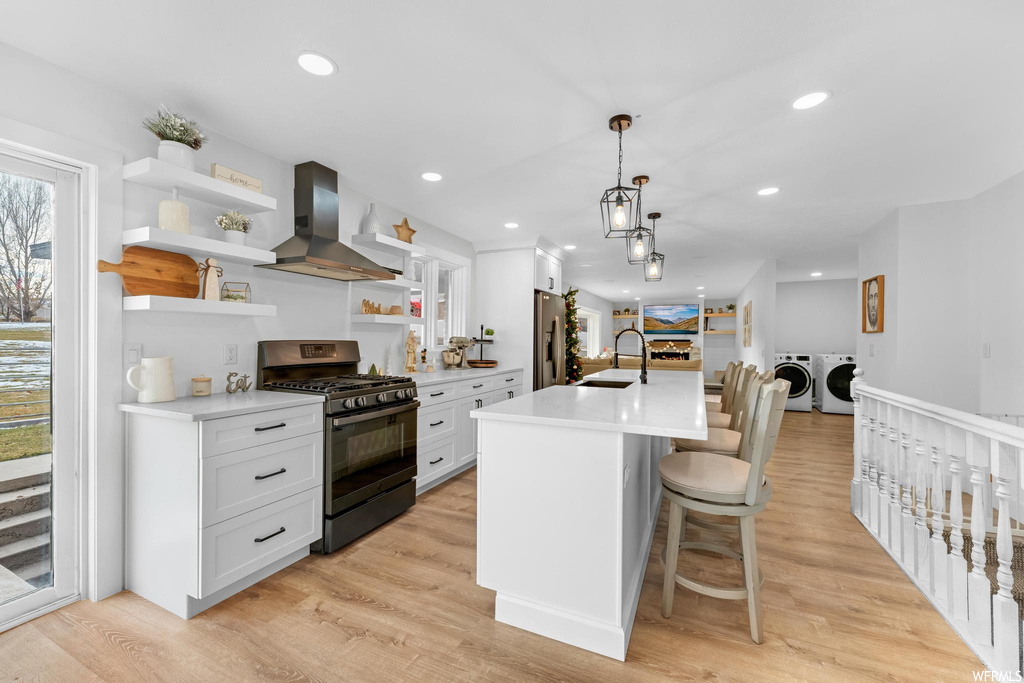 Kitchen featuring light hardwood / wood-style flooring, black gas range oven, wall chimney exhaust hood, washer and clothes dryer, and white cabinetry