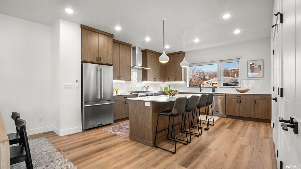 Kitchen with hanging light fixtures, light hardwood / wood-style floors, wall chimney range hood, appliances with stainless steel finishes, and a kitchen island