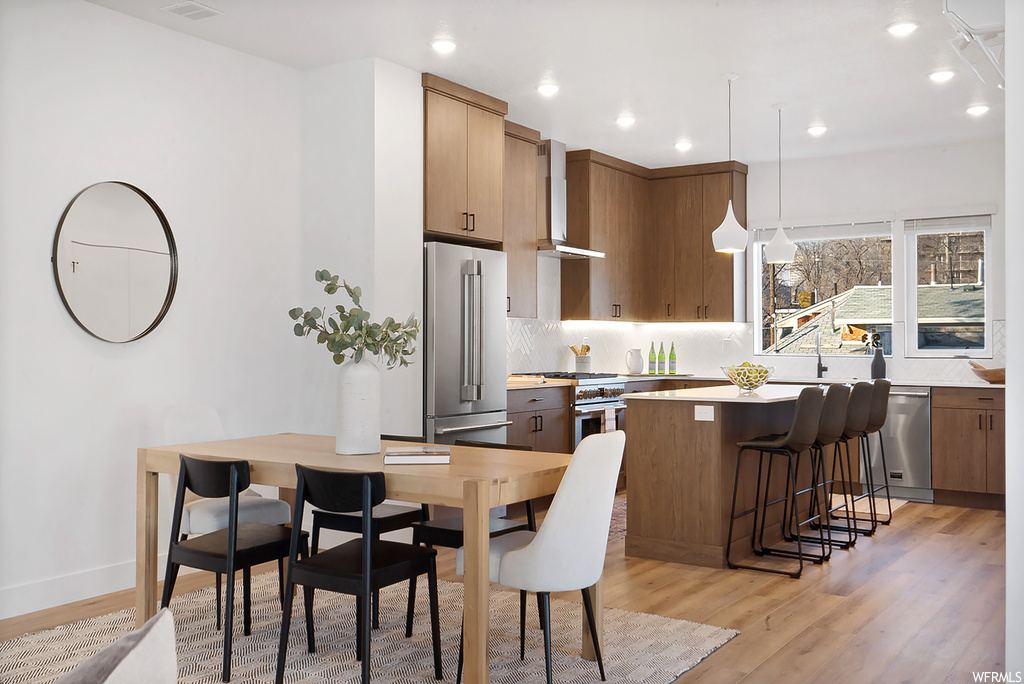 Kitchen with wall chimney range hood, hanging light fixtures, stainless steel appliances, light wood-type flooring, and tasteful backsplash