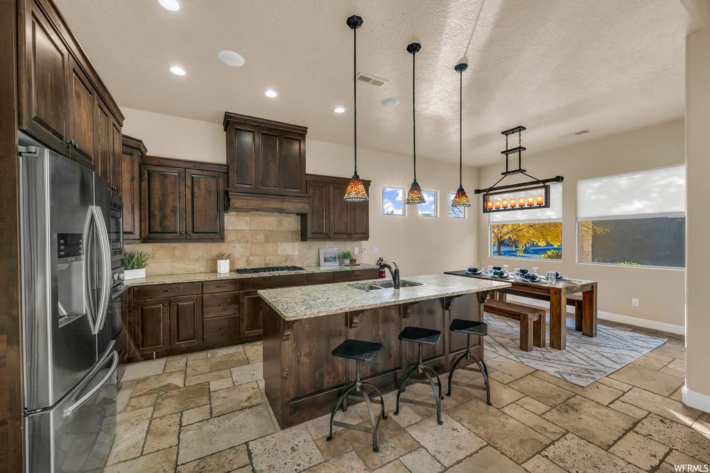 Kitchen with light stone counters, a center island with sink, hanging light fixtures, stainless steel appliances, and tasteful backsplash