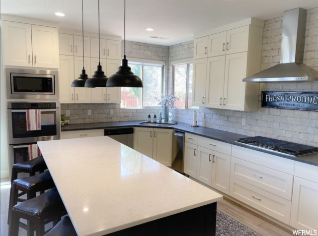 Kitchen featuring decorative light fixtures, appliances with stainless steel finishes, wall chimney range hood, and backsplash