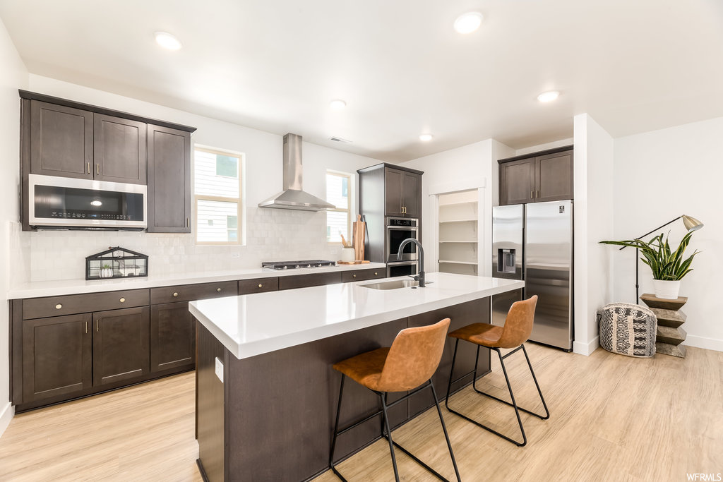 Kitchen featuring wall chimney exhaust hood, sink, appliances with stainless steel finishes, a kitchen island with sink, and light wood-type flooring
