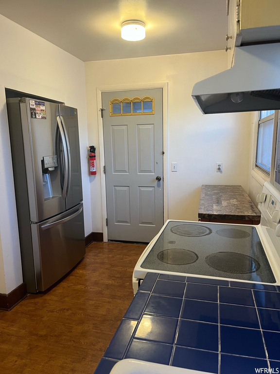 Kitchen with dark wood-type flooring, wall chimney exhaust hood, tile counters, and stainless steel fridge with ice dispenser