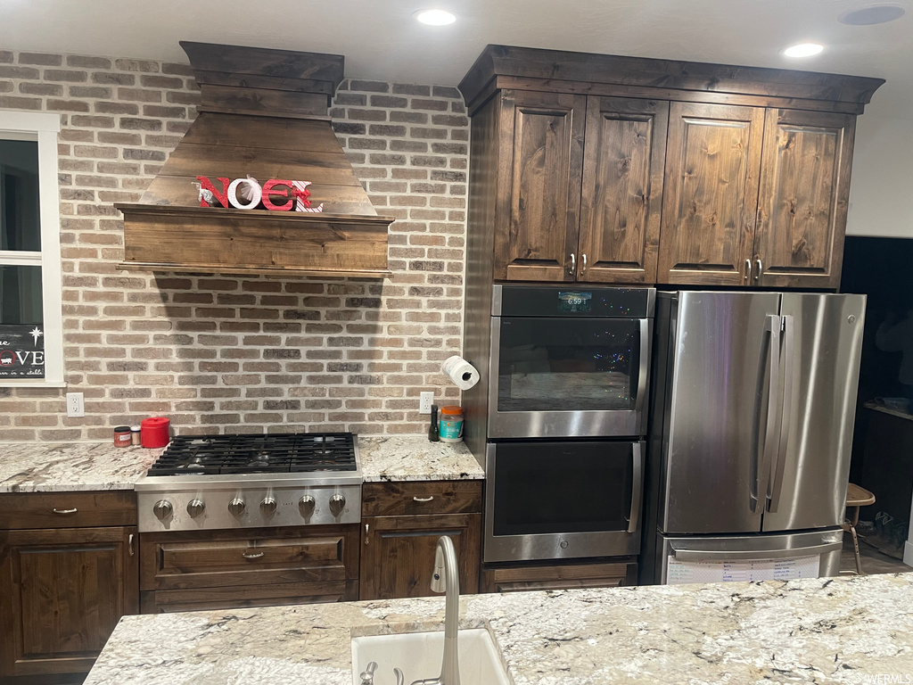 Kitchen with sink, appliances with stainless steel finishes, dark brown cabinetry, light stone countertops, and brick wall