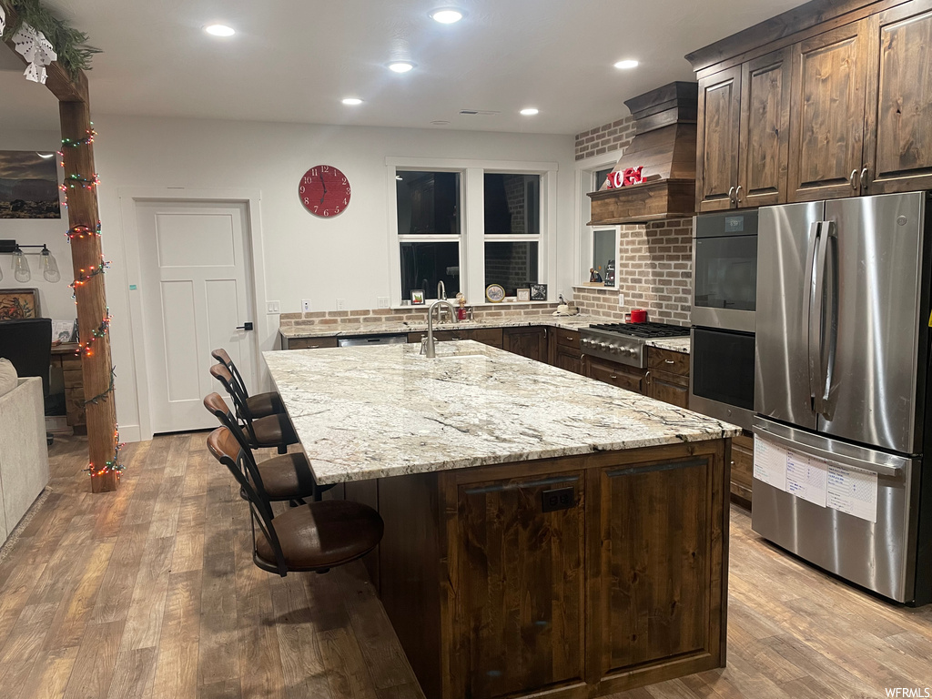 Kitchen with premium range hood, appliances with stainless steel finishes, light stone countertops, backsplash, and light hardwood / wood-style flooring