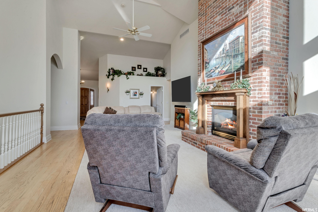 Living room featuring a fireplace, high vaulted ceiling, light hardwood / wood-style floors, ceiling fan, and brick wall