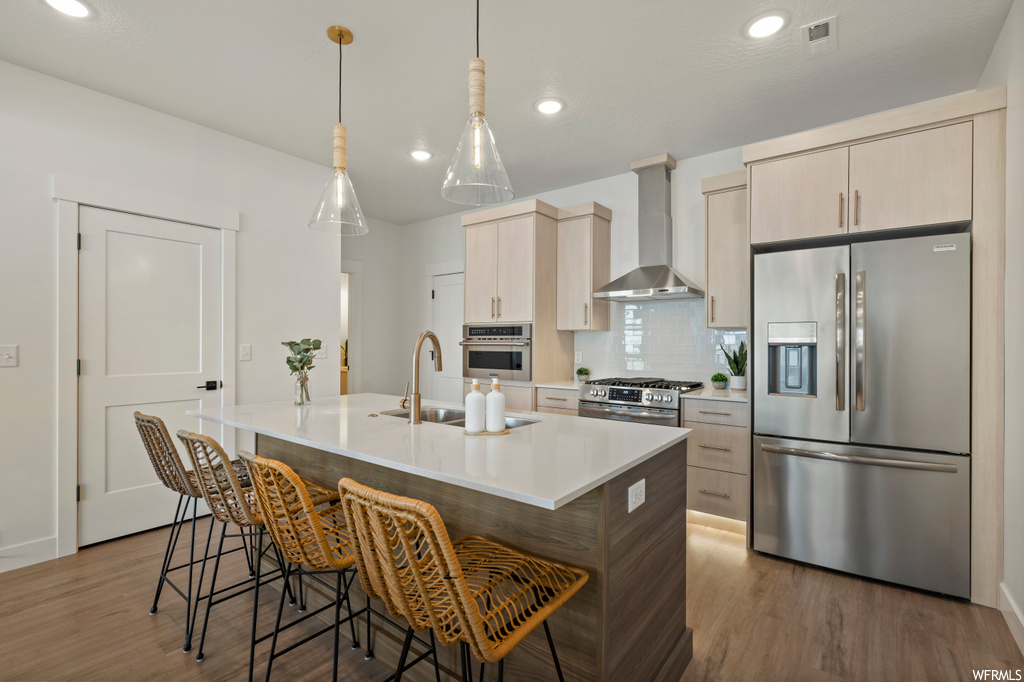 Kitchen featuring sink, wall chimney range hood, a kitchen island with sink, appliances with stainless steel finishes, and light wood-type flooring