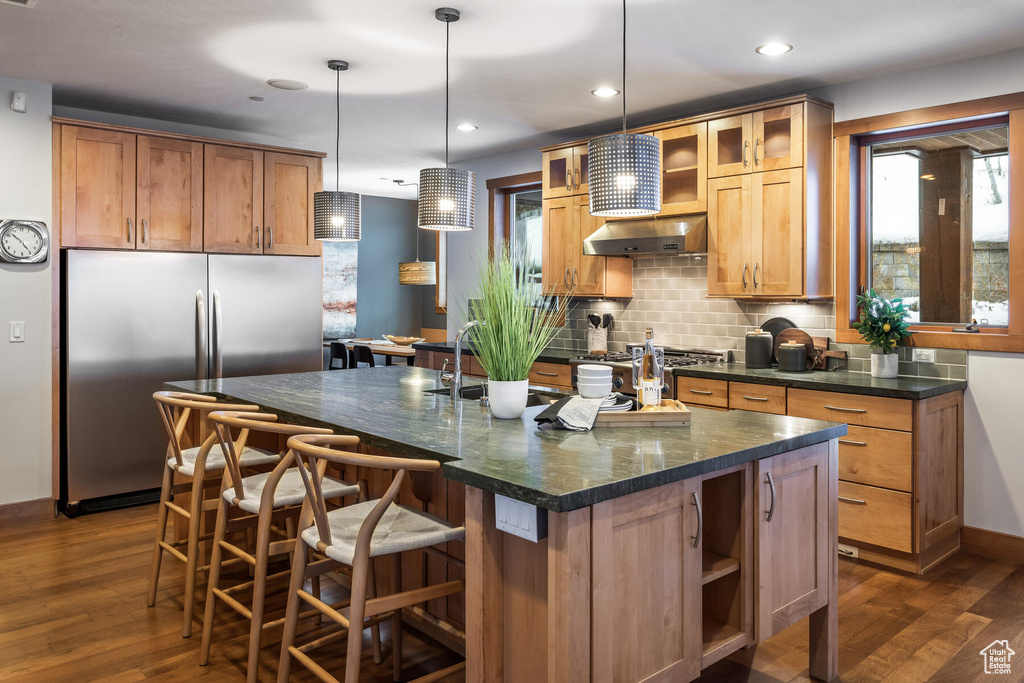 Kitchen with wall chimney range hood, dark wood-type flooring, stainless steel fridge, a center island with sink, and backsplash