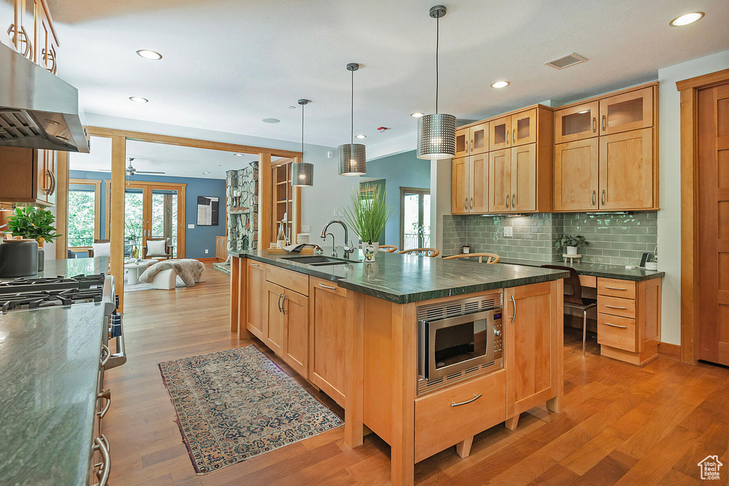 Kitchen with light hardwood / wood-style flooring, tasteful backsplash, stainless steel microwave, wall chimney exhaust hood, and sink