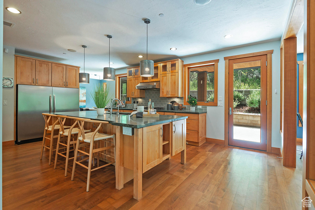 Kitchen with light hardwood / wood-style floors, a kitchen island with sink, backsplash, stainless steel refrigerator, and pendant lighting