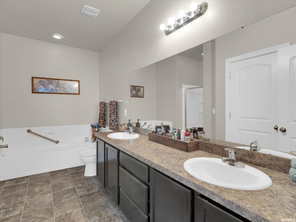 Bathroom featuring toilet, oversized vanity, double sink, tile floors, and a bath to relax in