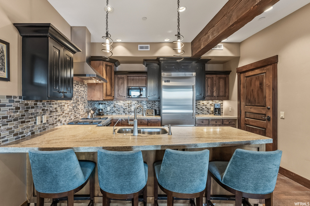 Kitchen featuring wall chimney exhaust hood, sink, light stone counters, built in appliances, and backsplash