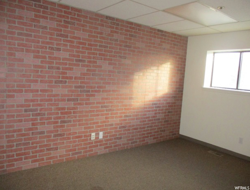 Empty room with dark carpet, a paneled ceiling, and brick wall