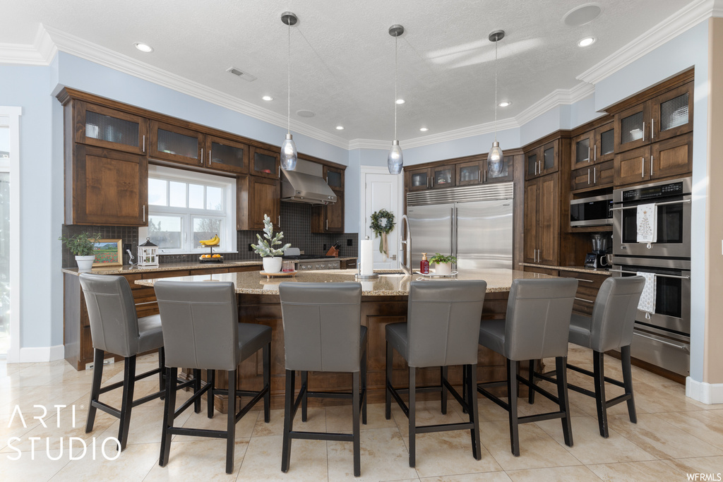 Kitchen with wall chimney exhaust hood, appliances with stainless steel finishes, pendant lighting, tasteful backsplash, and a center island with sink