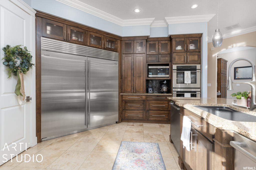 Kitchen featuring sink, built in appliances, light stone countertops, ornamental molding, and decorative light fixtures